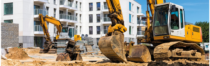 Zwei Bagger in Aktion im Vordergrund. Im Hintergrund ein dreigeschossiges Wohngebäude im Bau, weiß verputzt.